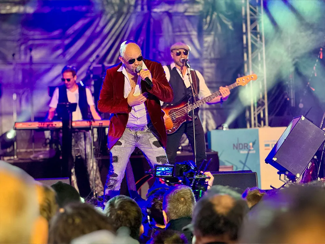 Der Sänger von Johnny Stone im roten Samtsakko singt ins Mikrofon, hinter ihm spielen Keyboarder und Bassist.