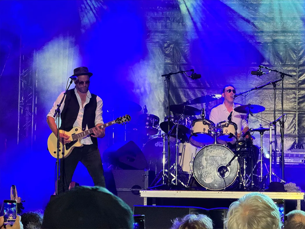Der Gitarrist und der Schlagzeuger von Johnny Stone in blauem Bühnenlicht, davor Zuschauer, die mit Handys filmen.
