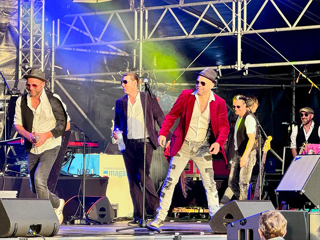 Johnny Stone tanzt und lacht auf der Bühne, der Sänger im roten Sakko steht in der Mitte, die Band um ihn herum.