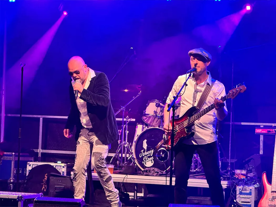 Der Sänger von Johnny Stone singt ins Mikrofon, neben ihm spielt der Bassist, im Hintergrund das Schlagzeug mit Bandlogo.